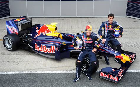 two men sitting on top of a red bull race car in front of another man