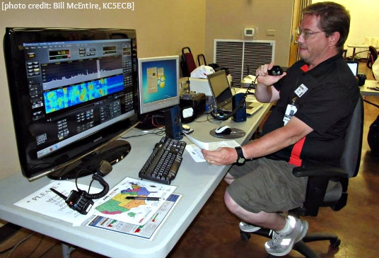 A man is seated at a computer talking on a hand held microphone indoors. The photo is credited to Bill McEntire KC5ECB.