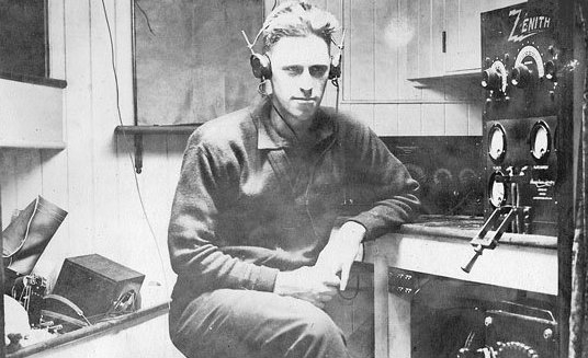 Black and white photo of man seated and is surrounded by radio equipment.